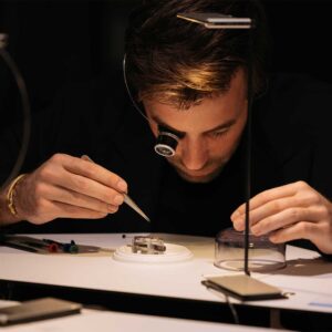 Uhrmacher beugt sich über eine Uhr, trägt eine Lupe und hat eine Pinzette in der Hand